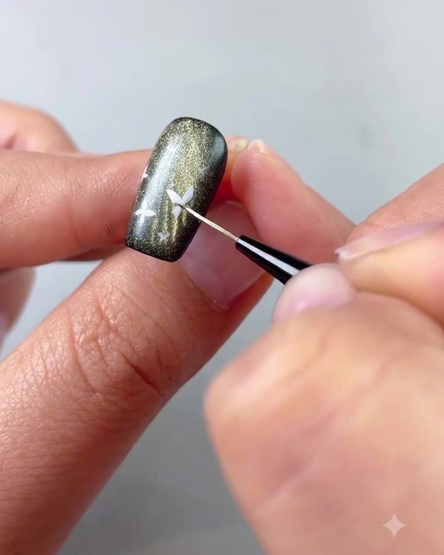 Primo piano di un pennello fine che applica gel bianco per dipingere un piccolo dettaglio di farfalla su ununghia scura cat-eye.