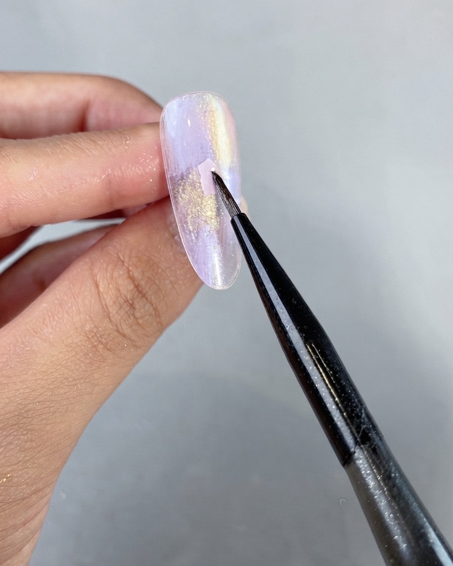 Un pennello liner fine per nail art che posiziona delicatamente un pezzo di conchiglia rosa sulla superficie di ununghia con gel perlato viola non polimerizzato.