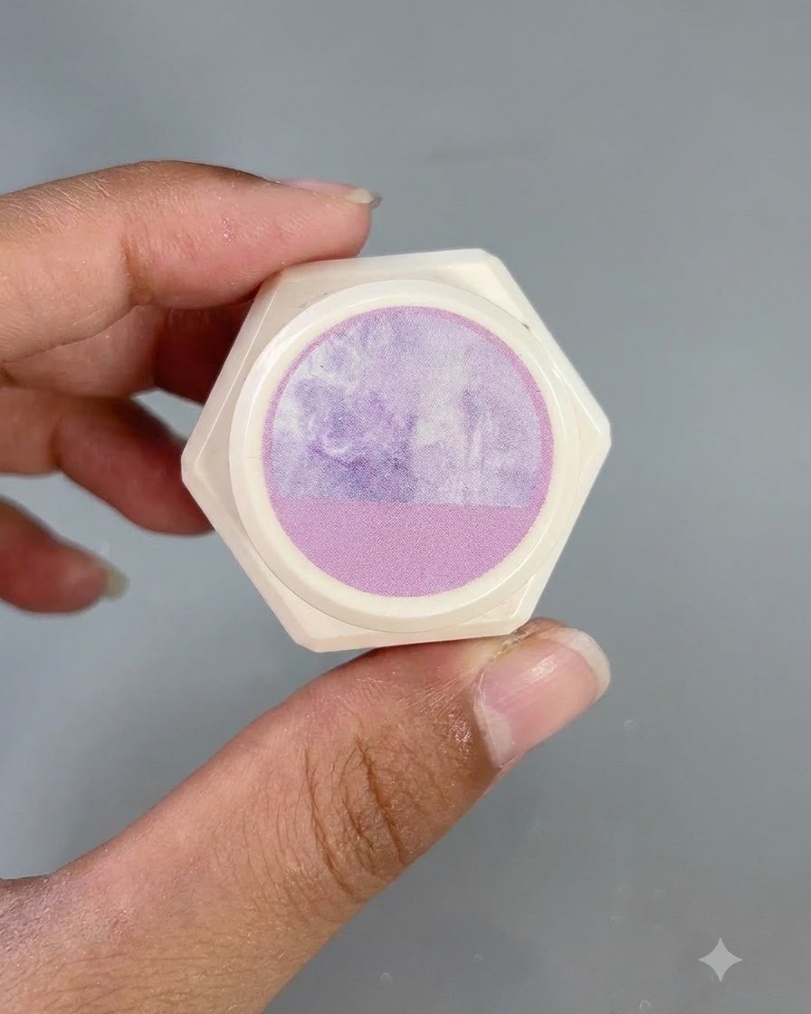 Un contenitore bianco esagonale con un adesivo con motivo acquarello viola sul coperchio, tenuto in alto dalle dita.