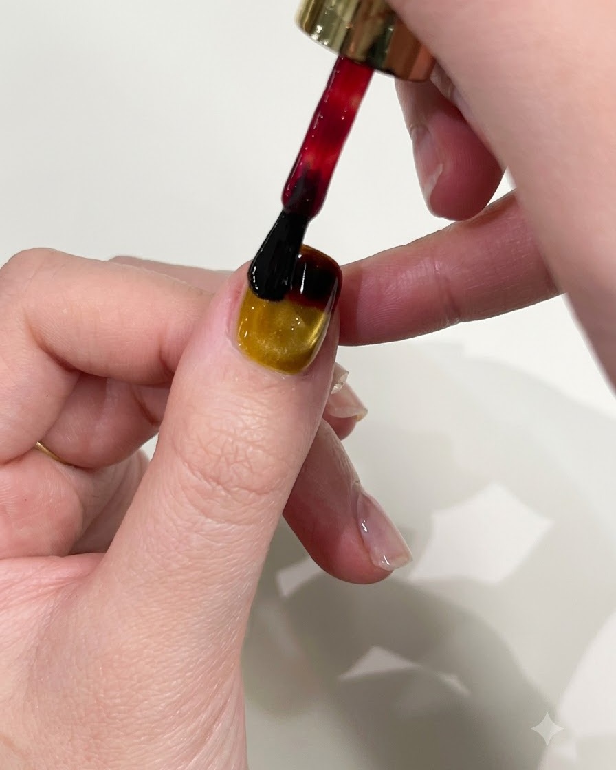 Un pennello per smalto che applica il gel rosso sul bordo superiore di ununghia con manicure cat-eye gialla.