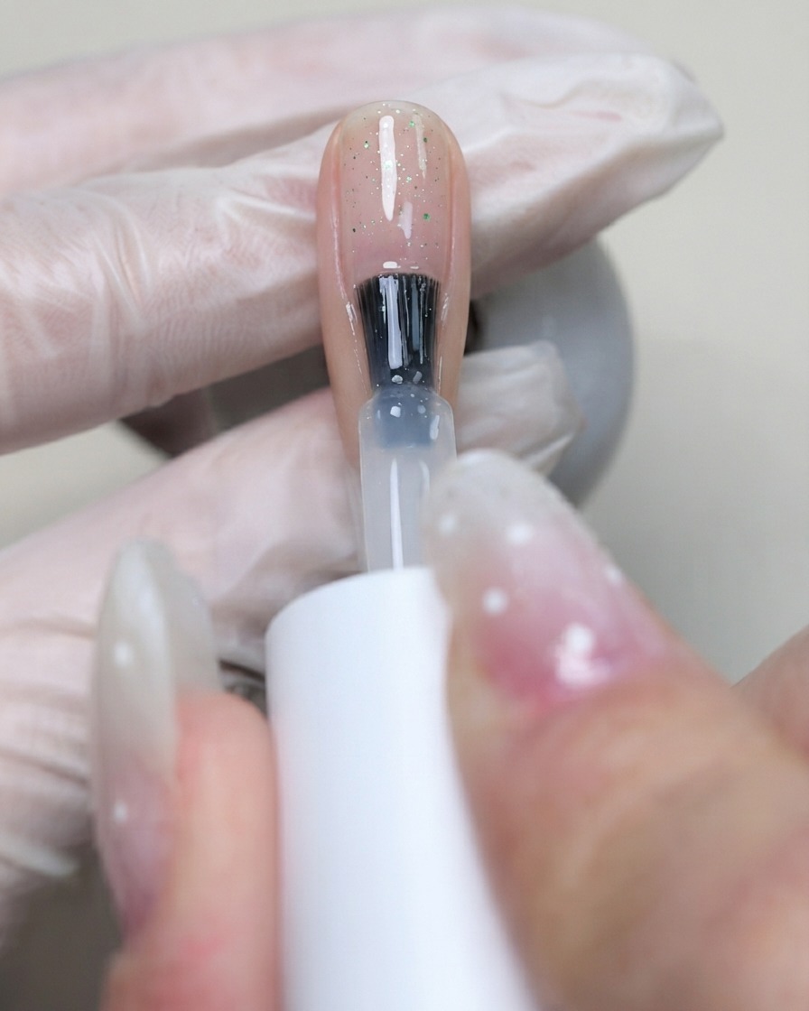 Un pennello che applica il base gel flessibile C7 trasparente sulla superficie dellunghia per bloccare le paillettes posizionate.