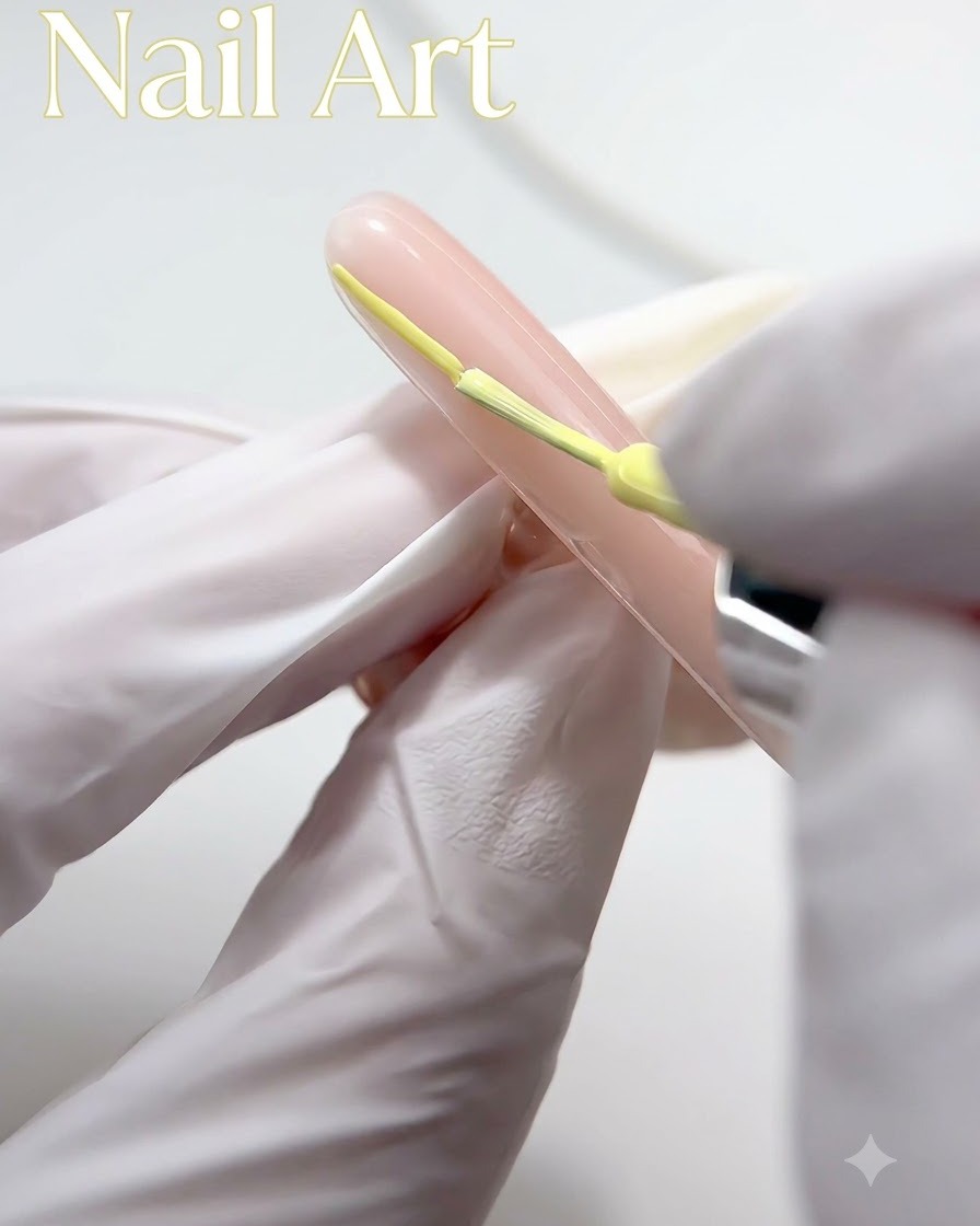 Una mano guantata che utilizza un pennello sottile per applicare lo smalto gel giallo lungo la punta di ununghia color nude.