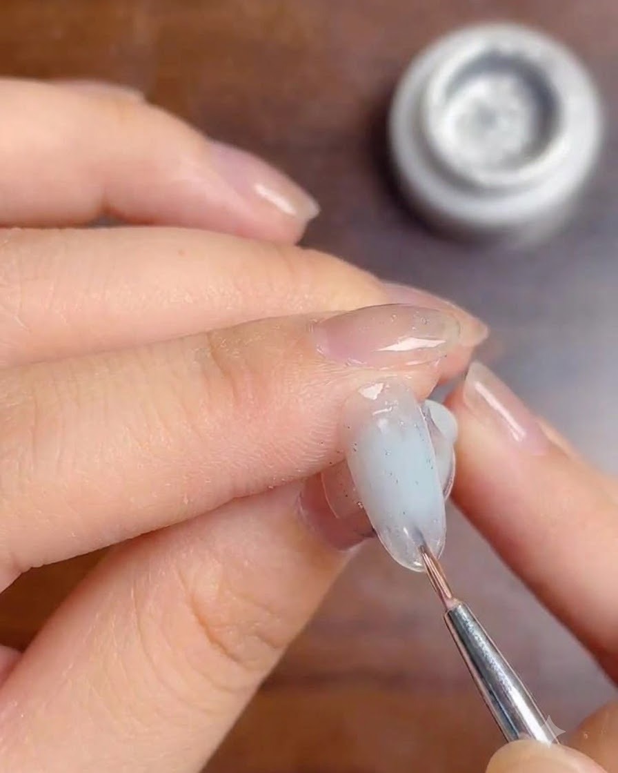 Un pennello fine che applica uno strato di gel top coat trasparente sopra le linee di glitter argento su ununghia sfumata bianco lattiginoso.