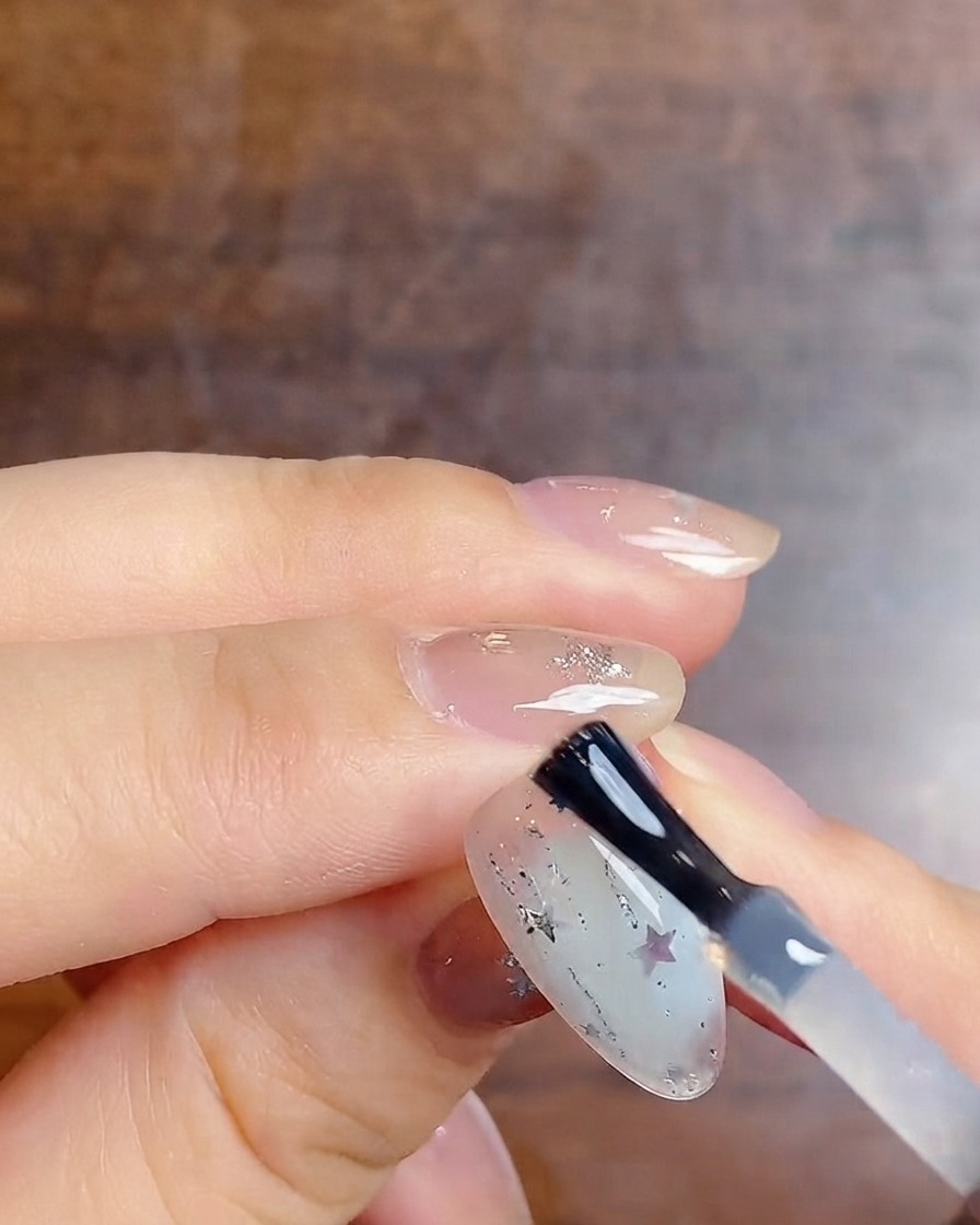 Un pennello per unghie che applica un top coat lucido finale su ununghia sfumata lattiginosa decorata con glitter argento e adesivi a stella.
