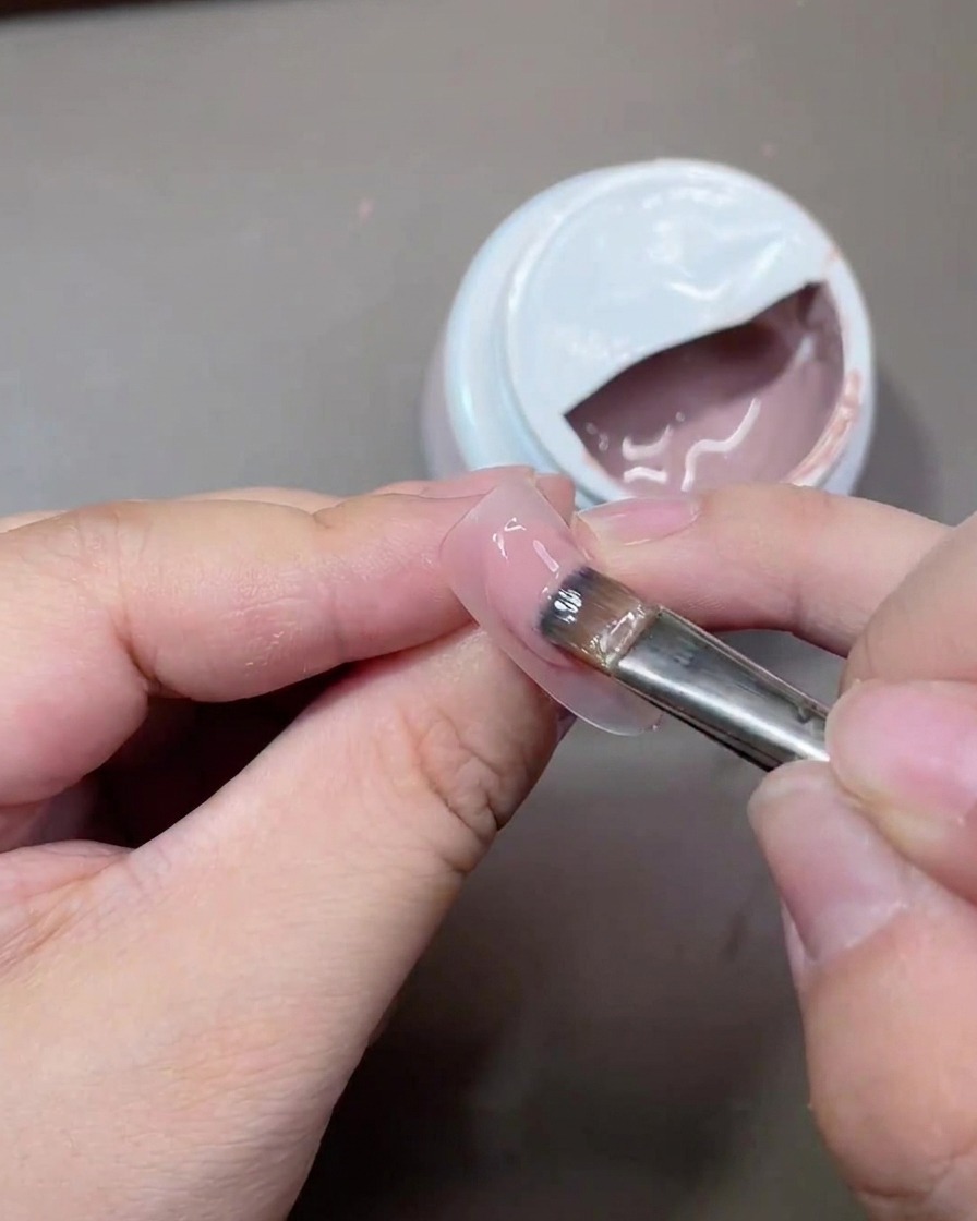 Un pennello da gel piatto che applica un sottile strato di smalto nude traslucido sulla superficie dellunghia.