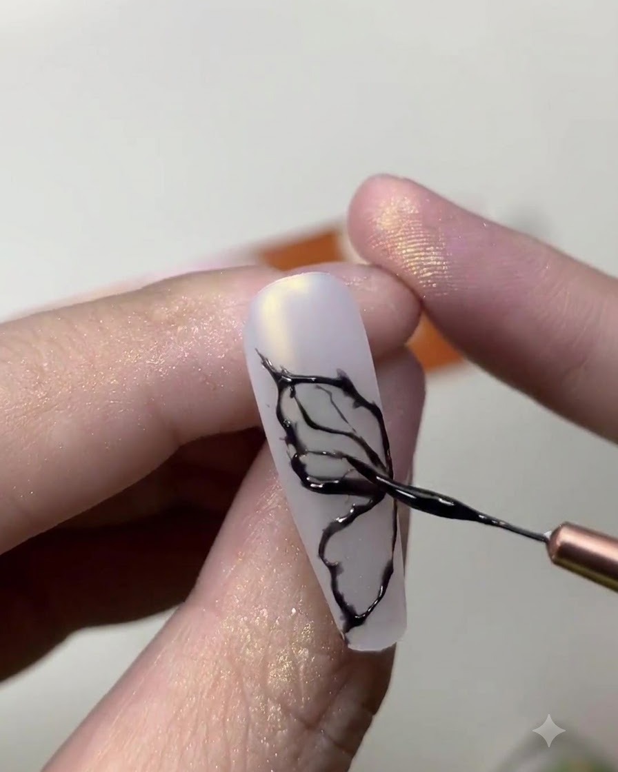 Un primo piano di una onicotecnica che utilizza un pennello sottile per disegnare delicate linee nere di ali di farfalla su ununghia bianco latte.