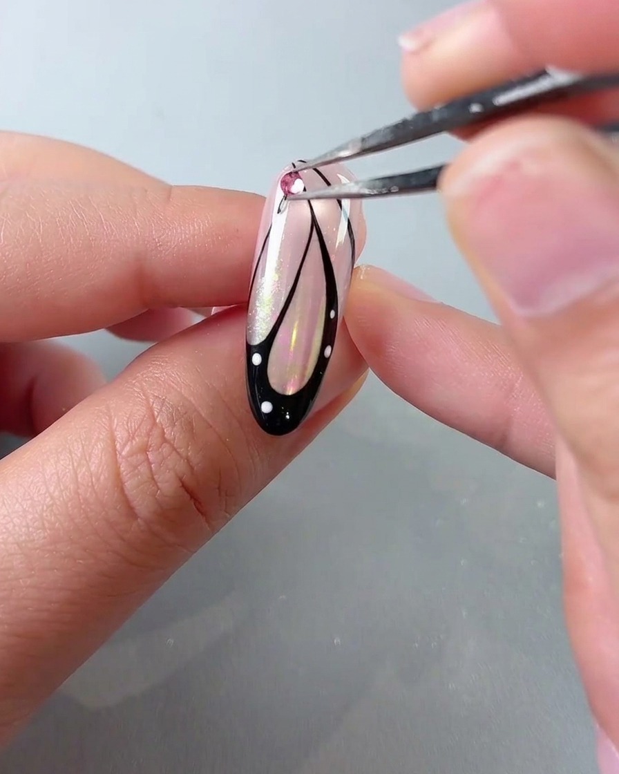 Un paio di pinzette che posizionano un rhinestone di cristallo rosa sopra un design per unghie con ali di farfalla.