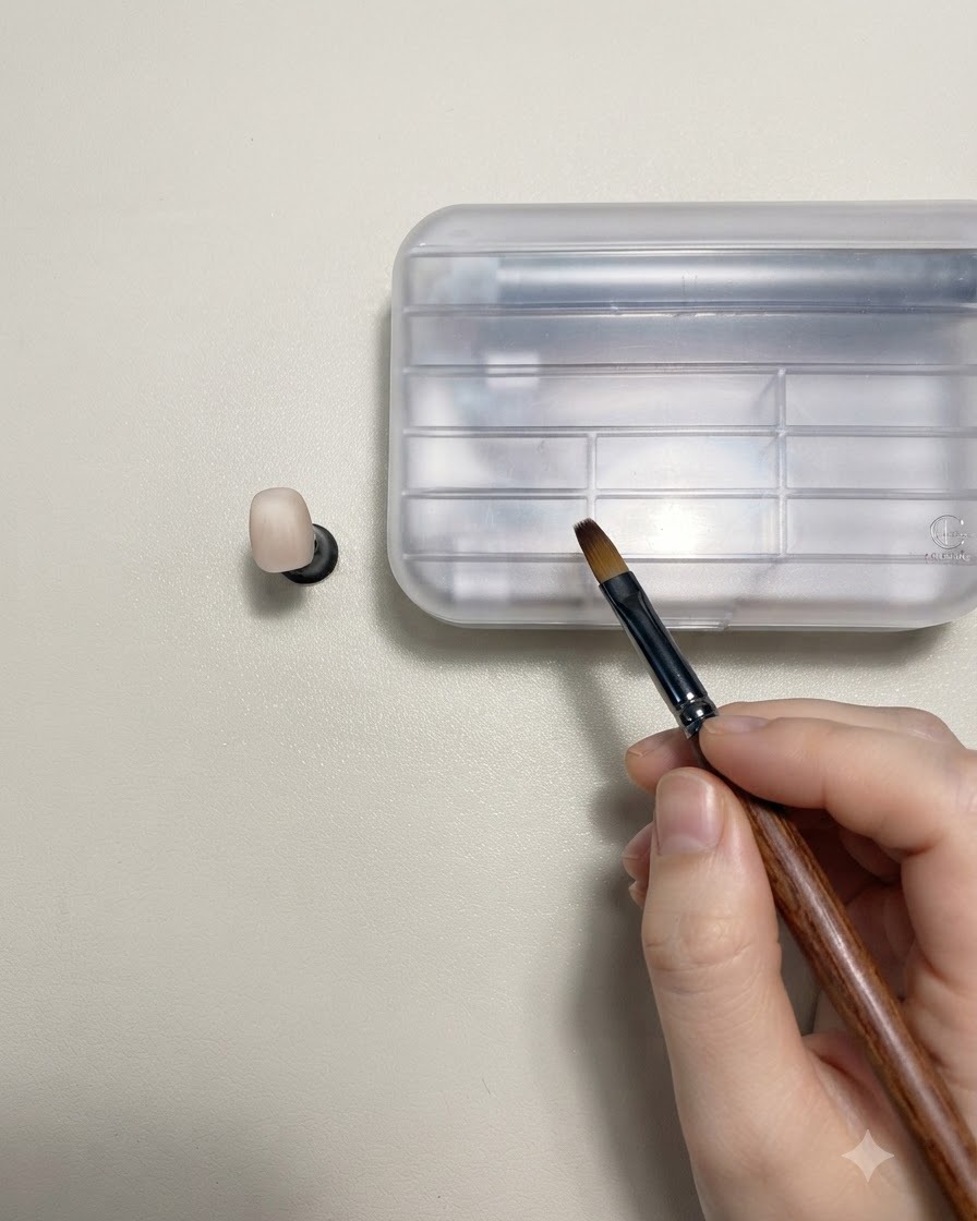 Una mano che tiene un pennello per nail art con manico in legno accanto a ununghia di prova color nude su un supporto.