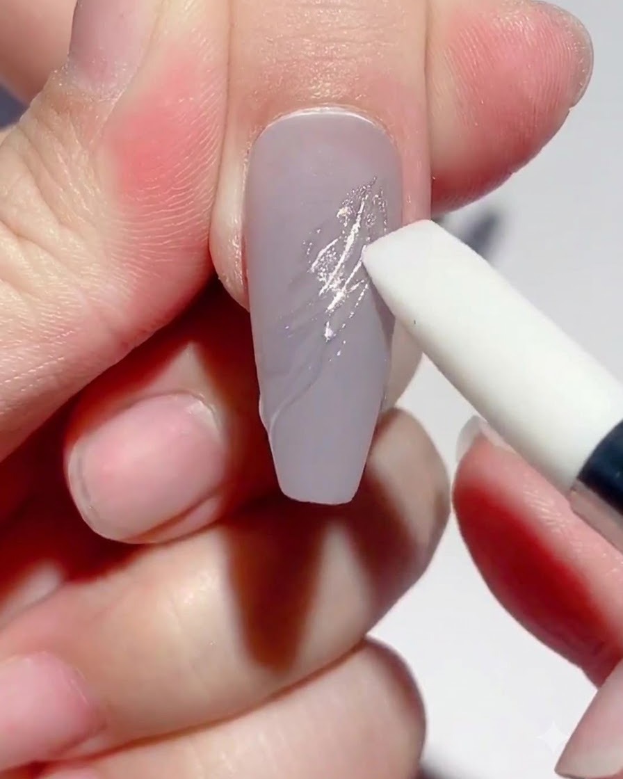 Uno strumento con penna in silicone che sfrega la polvere specchio argento sulle linee in gel polimerizzate su ununghia color tortora.