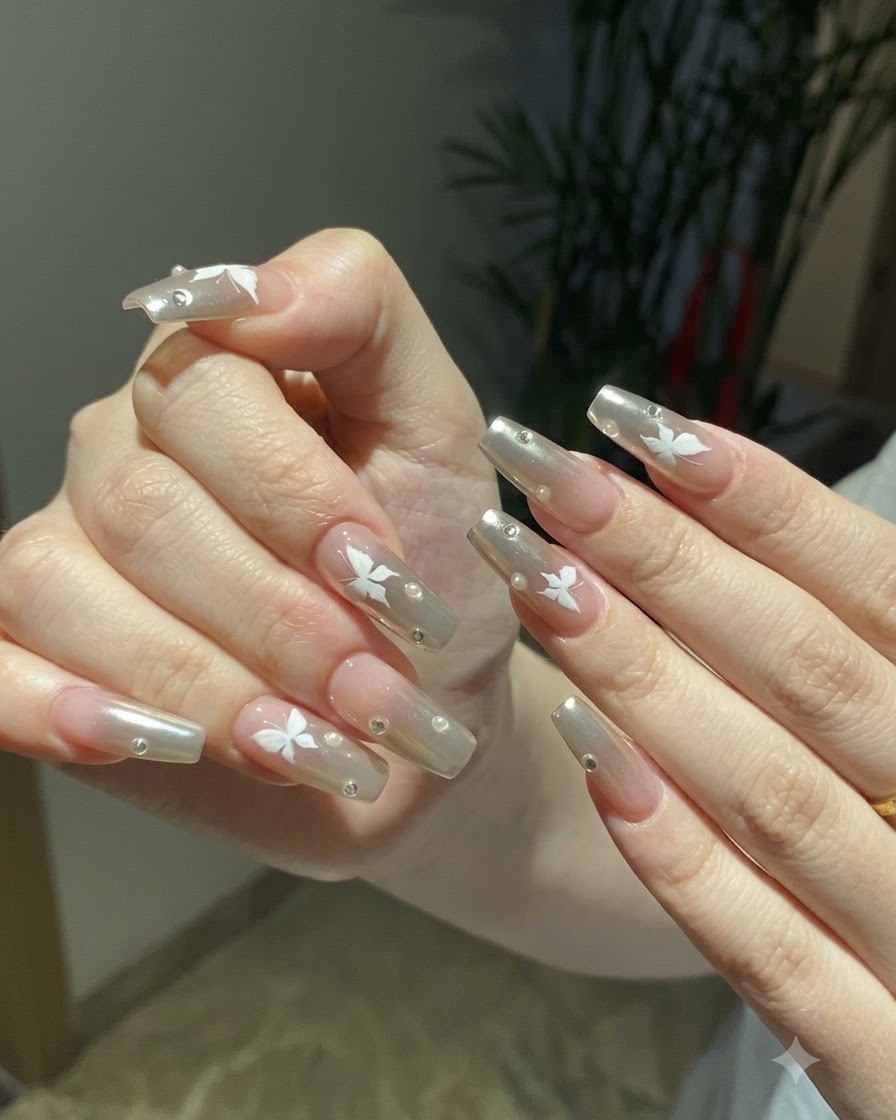 Due mani messe in posa insieme che mostrano la nail art completa con chrome argento e farfalle sotto una calda luce interna.