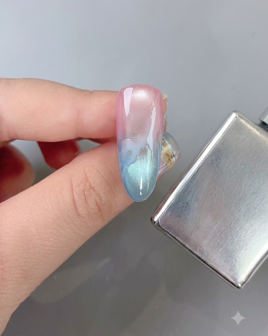 Un pennello per unghie che sfuma delicatamente i bordi di un motivo a nuvola bianca su ununghia sfumata multicolore accanto a uno stick magnetico.