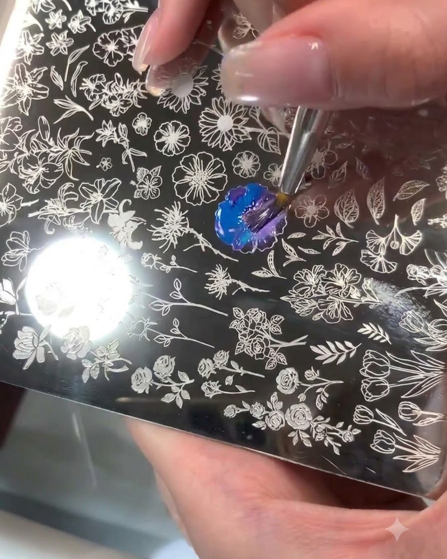 Una piastra per stamping in acciaio con un motivo floreale inciso in cui il gel blu e viola viene miscelato con un pennello.