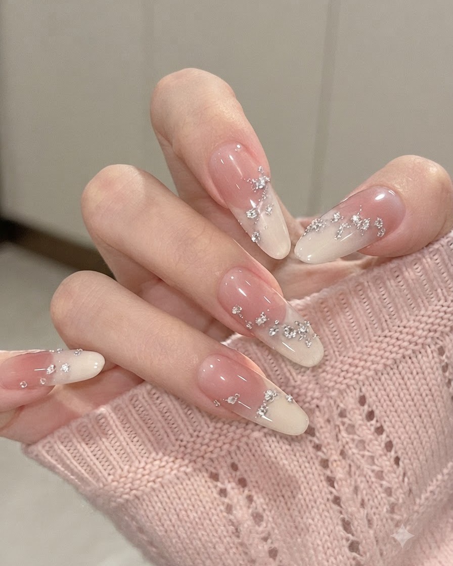 Primo piano di unghie a mandorla sfumate con dita leggermente piegate, che mettono in risalto le scaglie di foglia argento e i piccoli strass incorporati.