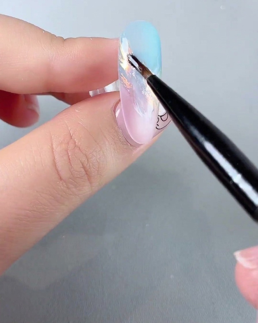 Un pennello fine per nail art che applica dettagli scintillanti in polvere glitter iridescente su ununghia sfumata pastello.