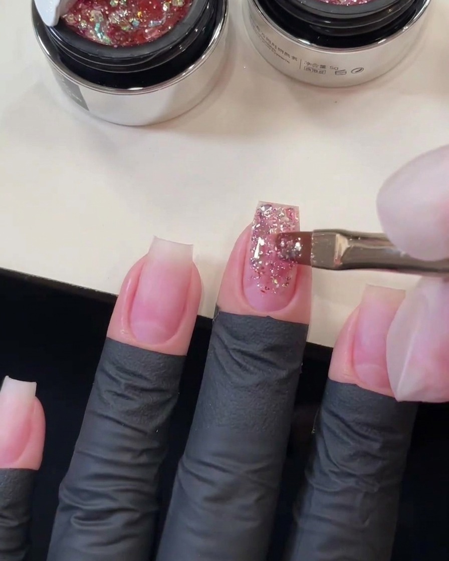Pennello piatto da gel che spazza le sequins rosa spesse lungo la lunghezza dellunghia del dito medio per formare un gradiente di glitter.