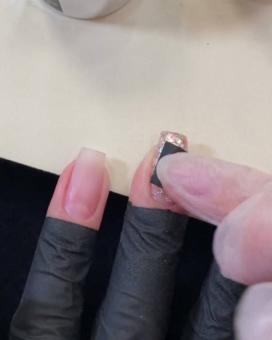 Un dito che preme un piccolo adesivo quadrato nero con un ritaglio a forma di stella su ununghia scintillante e con sequins.