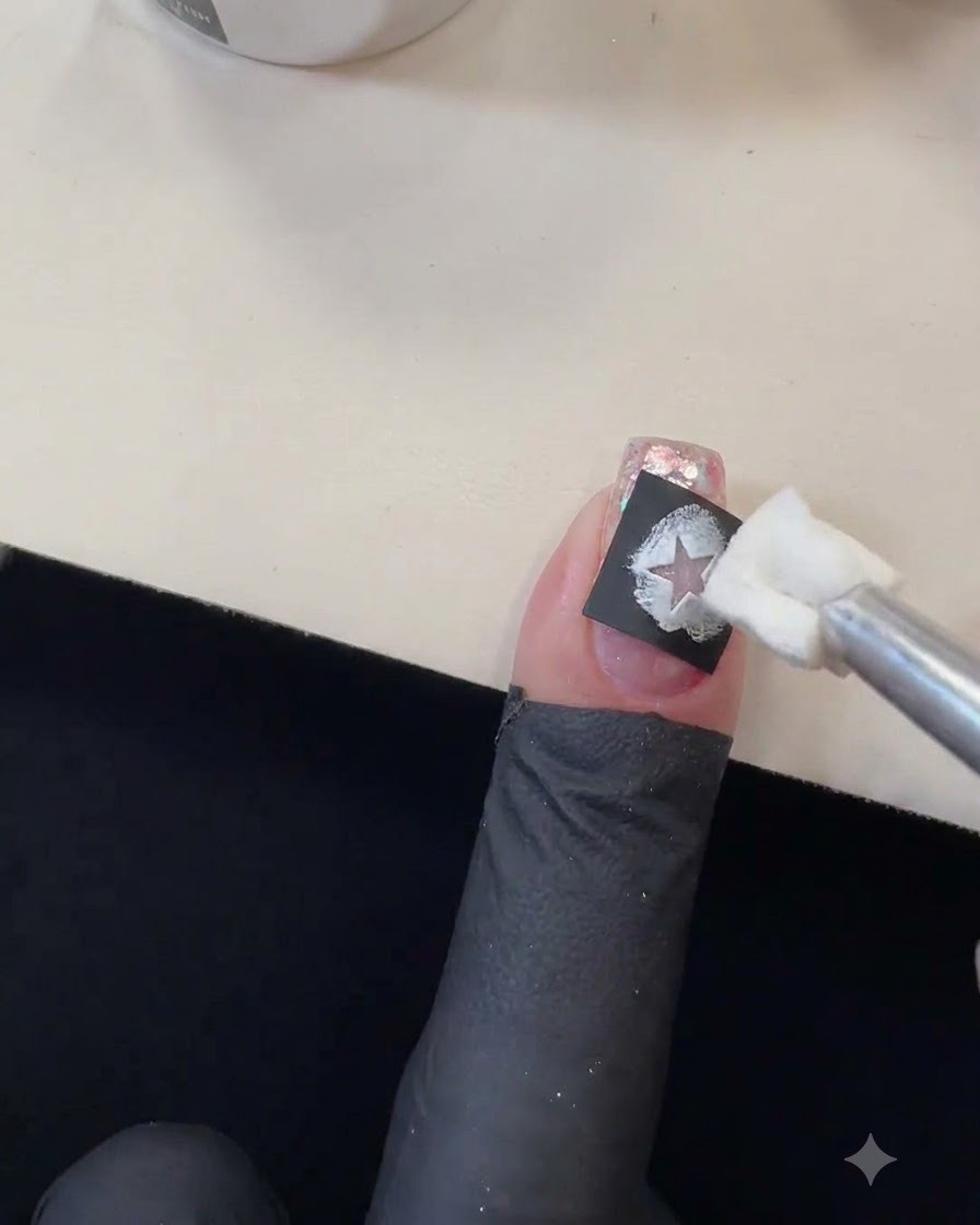 Un applicatore in spugna che tampona smalto gel bianco nel ritaglio di uno stencil a stella nero su ununghia glitterata.