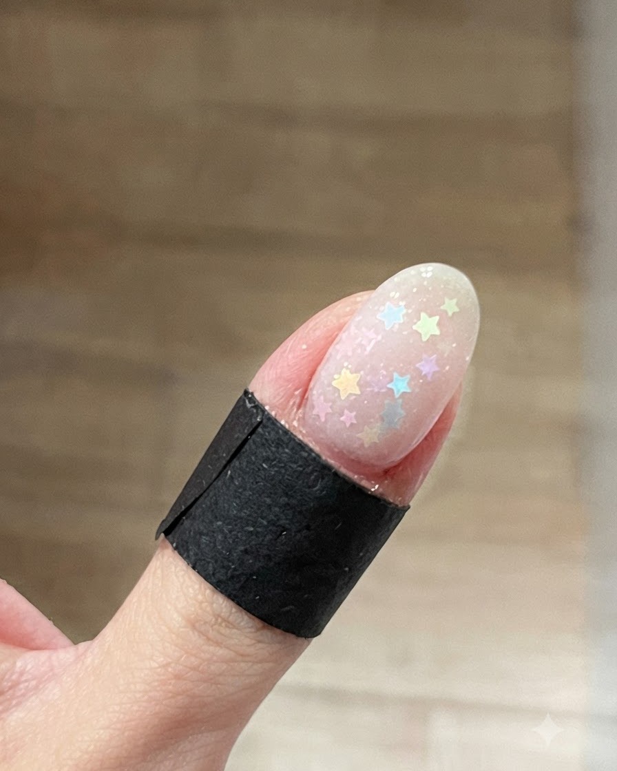 Un primo piano di ununghia ovale con glitter a stella multicolore sparsi su una base gel trasparente e bagnata.