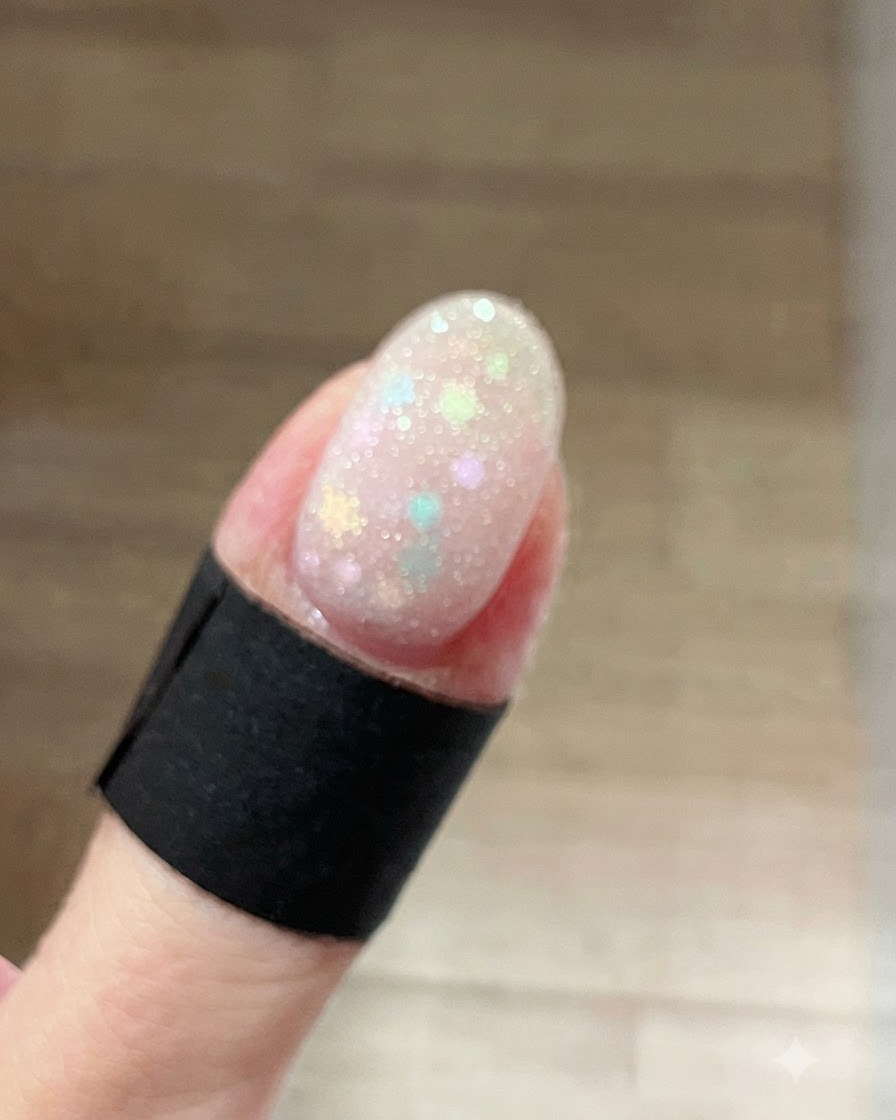 Un primo piano di ununghia ricoperta da un gel shimmer bianco ghiaccio, bagnato e scintillante, applicato sopra i glitter a stella pastello.