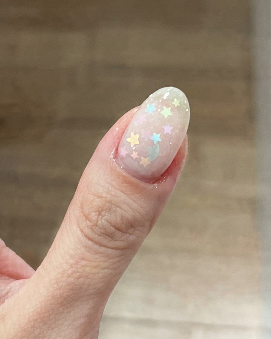 Un primo piano di ununghia del pollice caratterizzata da una base rosa lattiginoso con colorati glitter a stella pastello posizionati sopra.
