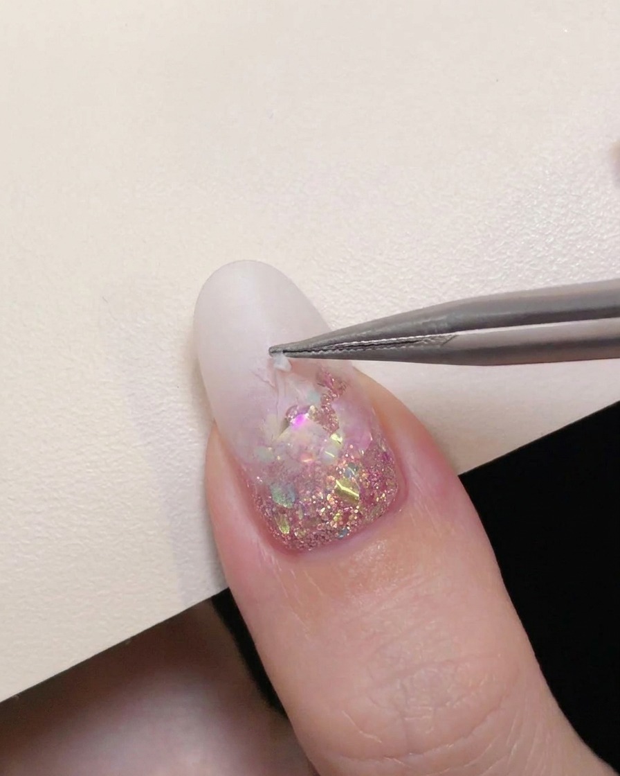 Le pinzette di precisione staccano con cura un adesivo a stella cava da ununghia con sfumatura bianco latte per rivelare i glitter rosa sottostanti.