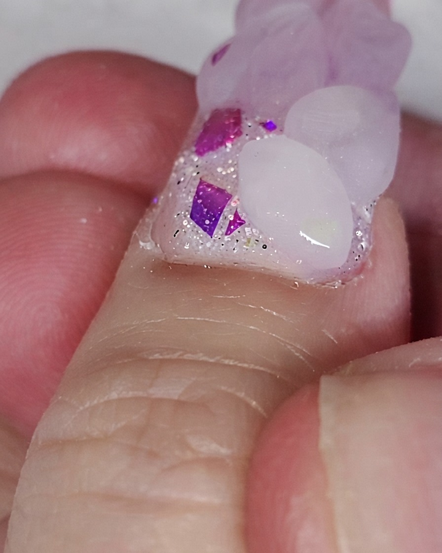 Un pennello da unghie posiziona una perla acrilica rosa su una base di unghie glitterata per formare la forma di un cuore.