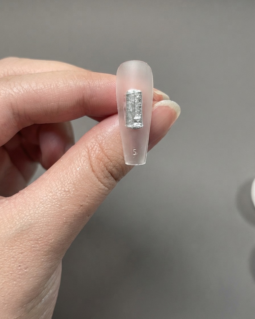 Un pezzo di extension per unghie trasparente tenuto tra le dita che mostra una pennellata nera o argentata applicata direttamente senza una base.