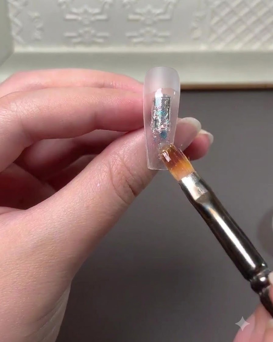 Un pennello per nail art che applica scaglie di glitter multicolore e polvere high gloss al centro di unextension per unghie trasparente.
