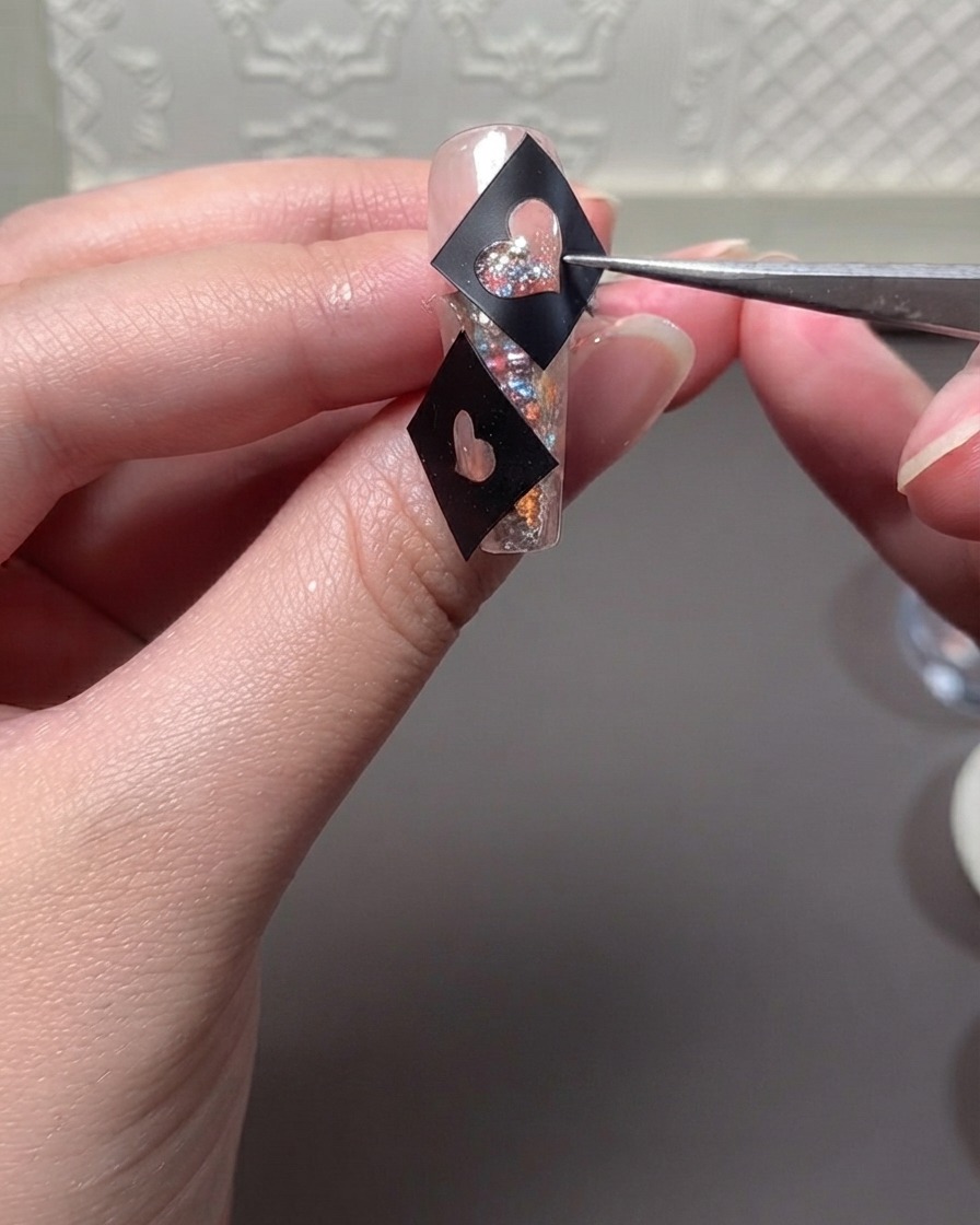 Un paio di pinzette che tengono un modello di stencil a cuore nero sopra ununghia piena di glitter mentre applicano il gel.