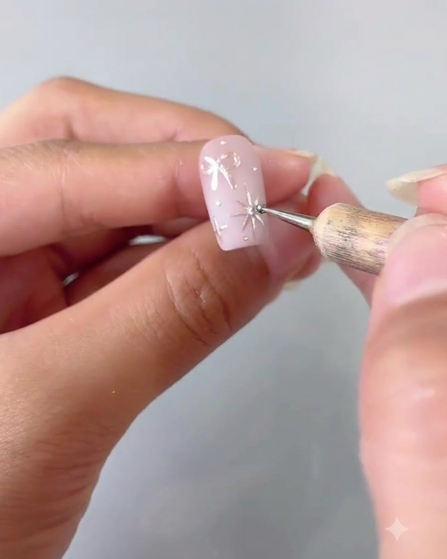Primo piano che mostra uno strumento dotting che posiziona con cura un minuscolo cristallo al centro di una stella argentata a quattro punte dipinta su ununghia rosa.