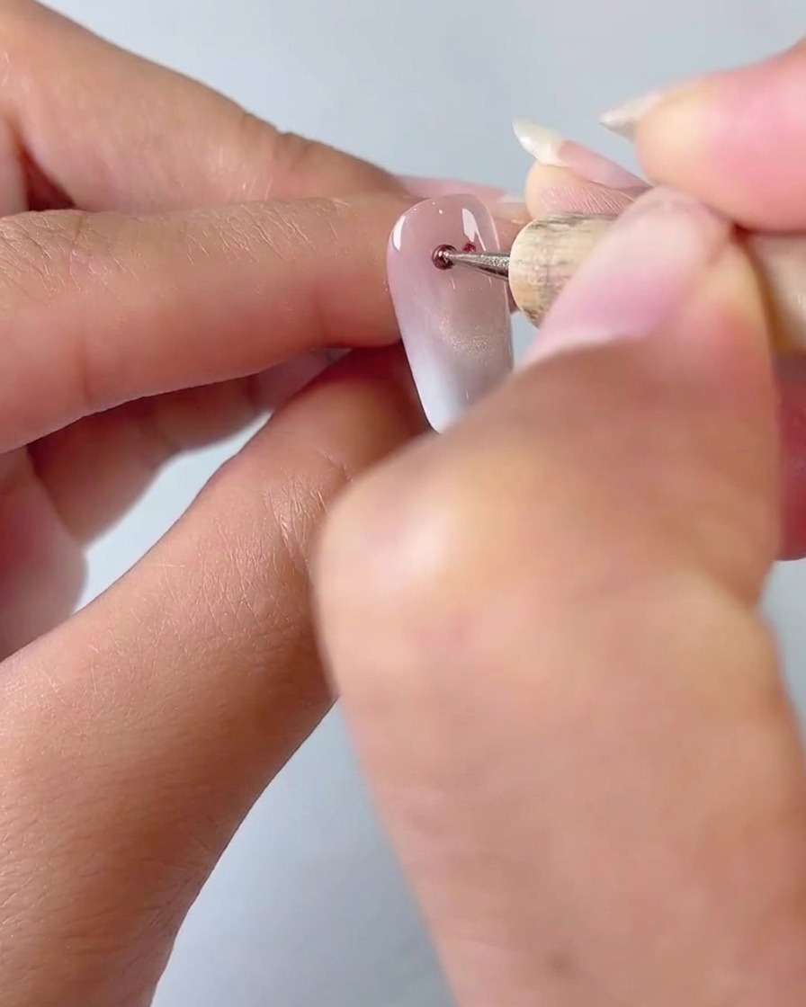Primo piano di uno strumento dotting che applica due piccoli punti di gel cat eye rosso su una base per unghie con sfumatura lattiginosa.
