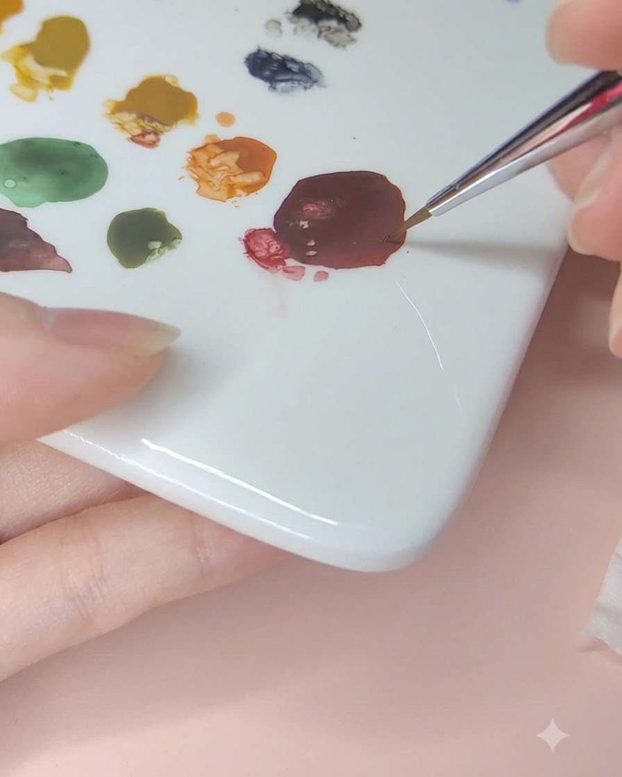 Un pennello fine da nail art che preleva pigmento per acquerello rosso scuro da una tavolozza di ceramica.