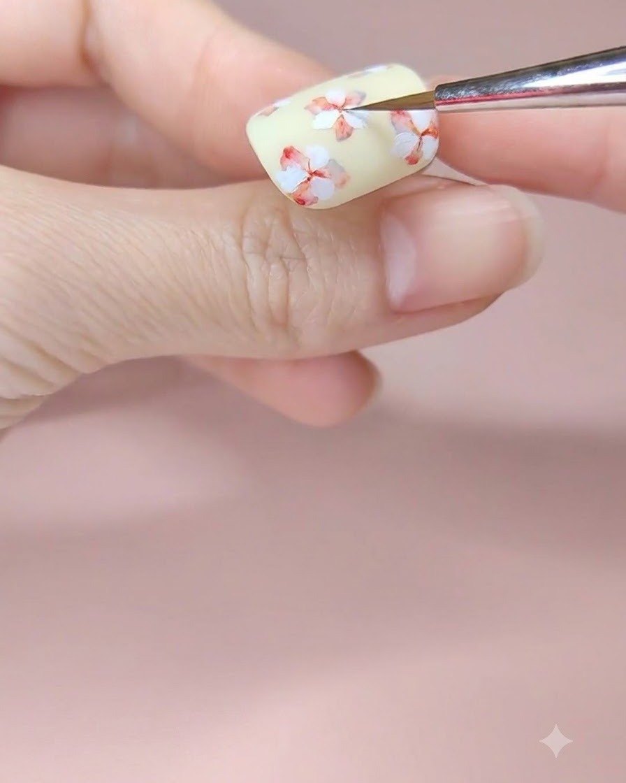 Un pennello fine da nail art che aggiunge un tocco di pigmento ad acquerello rosso al centro di un fiore dipinto di bianco su ununghia giallo pallido.