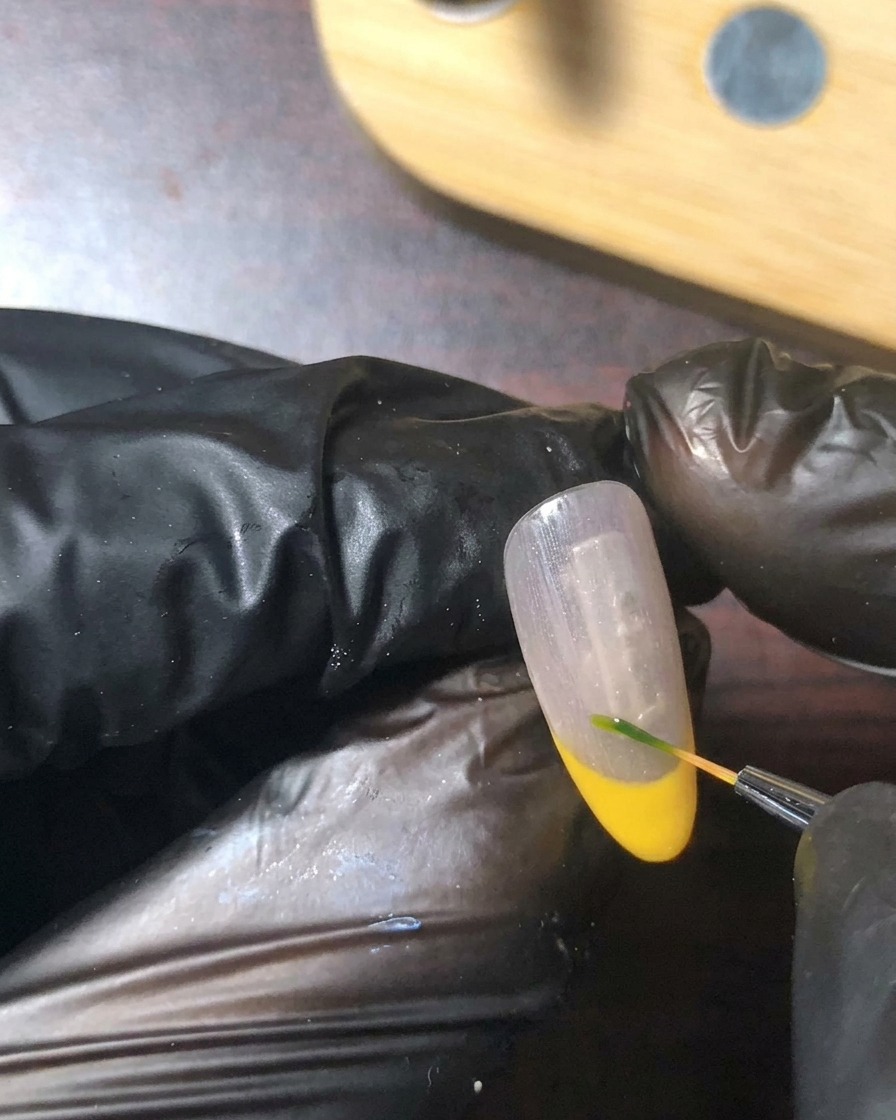 Un pennello di dettaglio fine che dipinge un minuscolo stelo verde e una foglia vicino alla linea del sorriso dellunghia.