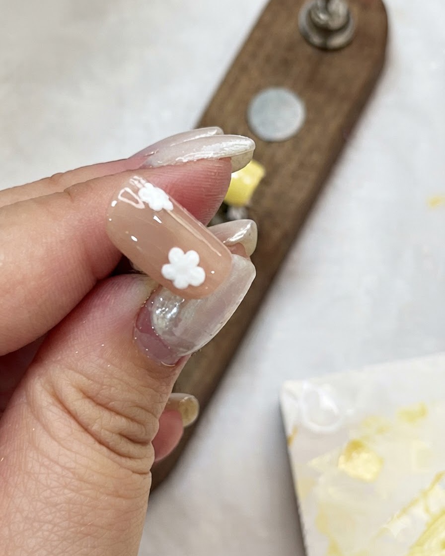 Una mano che tiene una tip per unghie color nudo mentre aggiunge petali a punti bianchi per creare fiori.