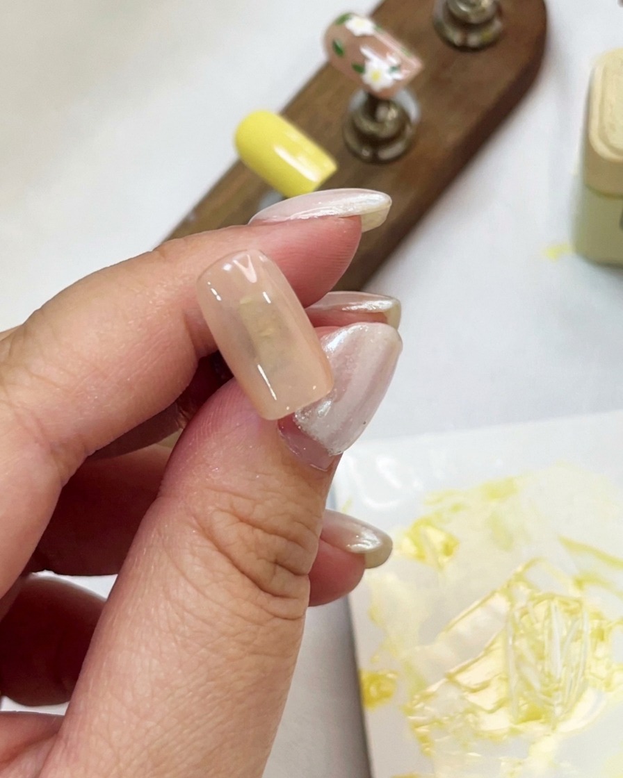 Primo piano di una mano che tiene una tip per unghie color nudo mentre applica il gel cat-eye brillante con un pennello.
