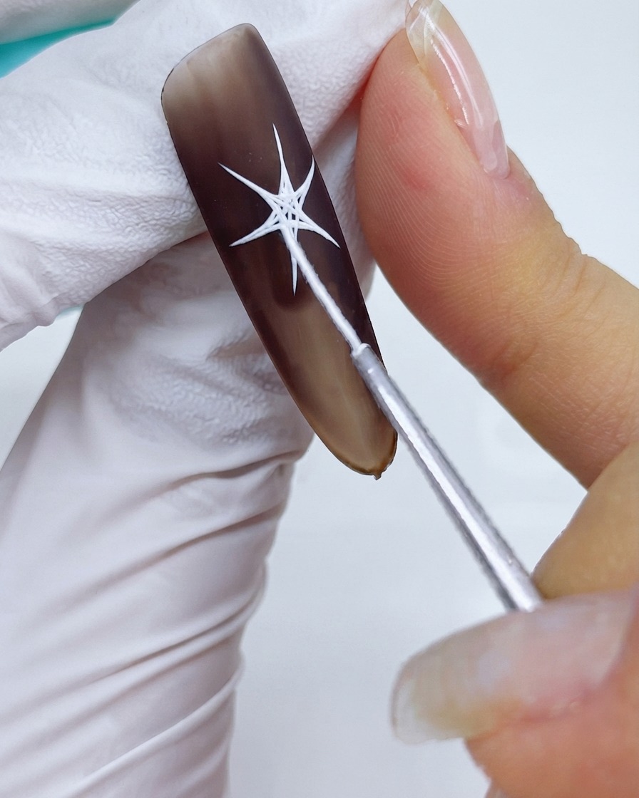 Primo piano di un pennello per dettagli che collega le punte e riempie le sezioni centrali di un design a stella bianco su una base gel marrone opaca.