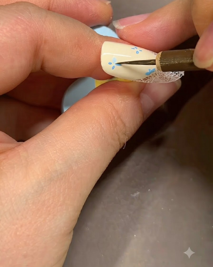 Un pennello fine per nail art che aggiunge dettagli blu al centro di un fiore dipinto su ununghia beige.
