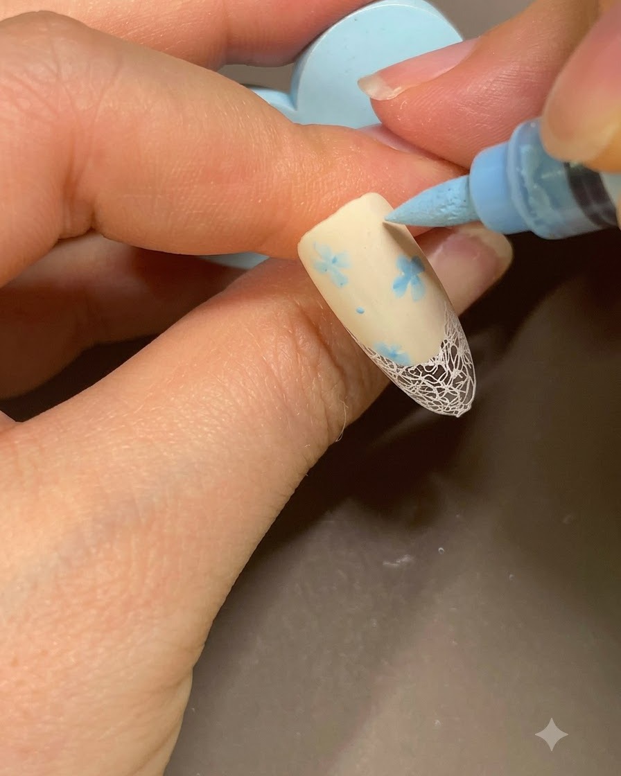 Un pennarello per nail art blu che aggiunge piccoli punti finali accanto a un pattern floreale su french manicure beige.