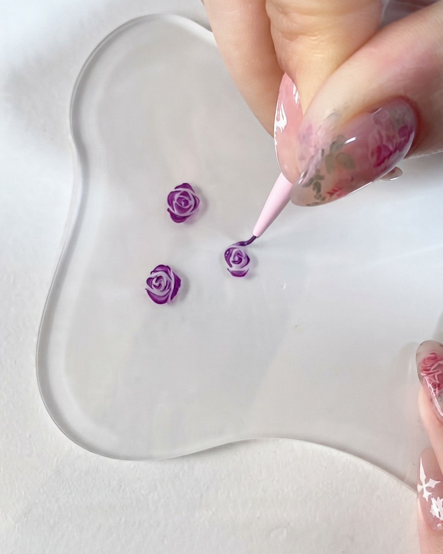 Un primo piano di una mano che regge un pennello da dettaglio fine, intento a dipingere meticolosamente accenti viola scuro su un piccolo design di rosa 3D su una palette di vetro trasparente.