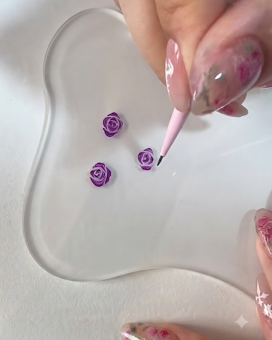 Un primo piano di una mano che usa un pennello da dettaglio rosa per rifinire i bordi di una rosa viola 3D dipinta su una palette trasparente.