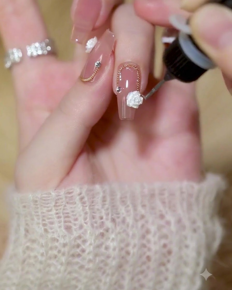 Un pinceau fin de détail appliquant du gel de construction transparent autour des bords dune rose blanche 3D et dune chaîne dorée sur un ongle poli.