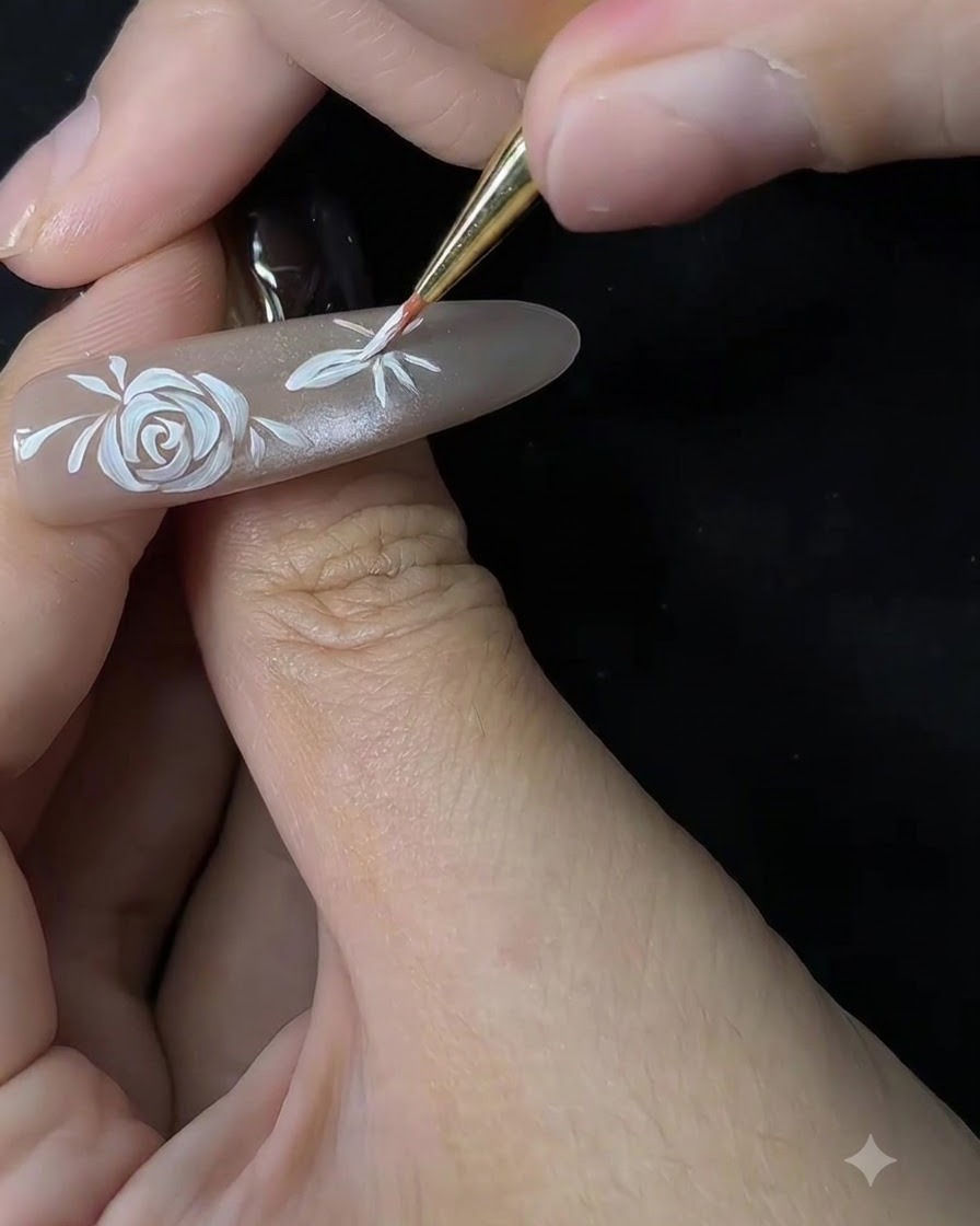 Primo piano di un pennello sottile che inizia a modellare i petali di un secondo fiore su unextension per unghie color nude.