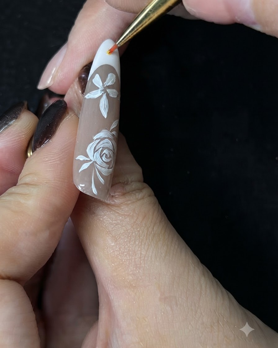 Primo piano di una mano che usa un pennello di precisione fine per dipingere piccoli tralci accento bianchi attorno a una rosa bianca dipinta a mano su ununghia in gel nude.