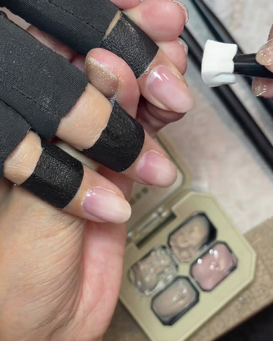 Uno strumento a spugnetta che tampona il gel di copertura nude latteo sul margine libero naturale di ununghia, con una tavolozza di gel sullo sfondo.
