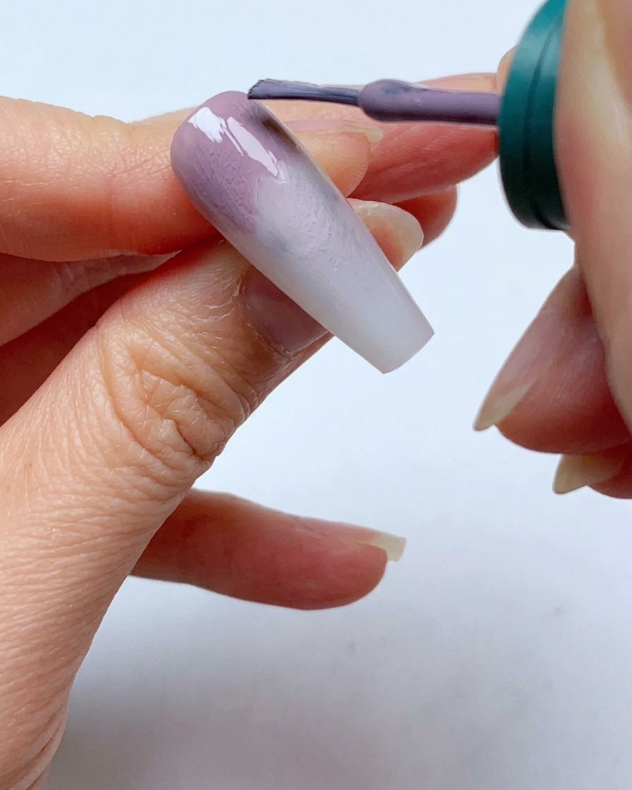 Un pennello per smalto applica un secondo strato di gel grigio-viola vicino alla cuticola di ununghia.