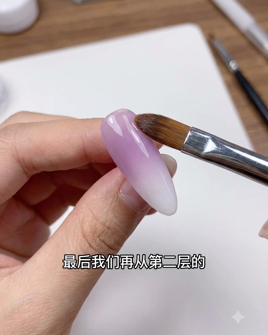 Un pennello per unghie pulito sfuma delicatamente la linea di transizione su ununghia con gradiente viola e bianco.