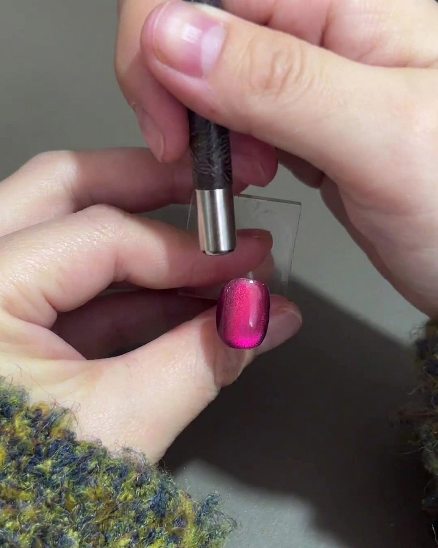 Una mano che tiene un magnete cilindrico sopra il bordo superiore di ununghia in gel viola per spingere il punto luce magnetico verso il basso.