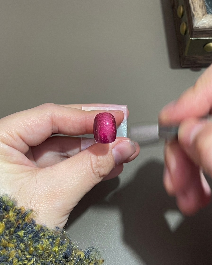 Una mano che tiene un piccolo magnete cilindrico vicino a ununghia in gel cat-eye viola.