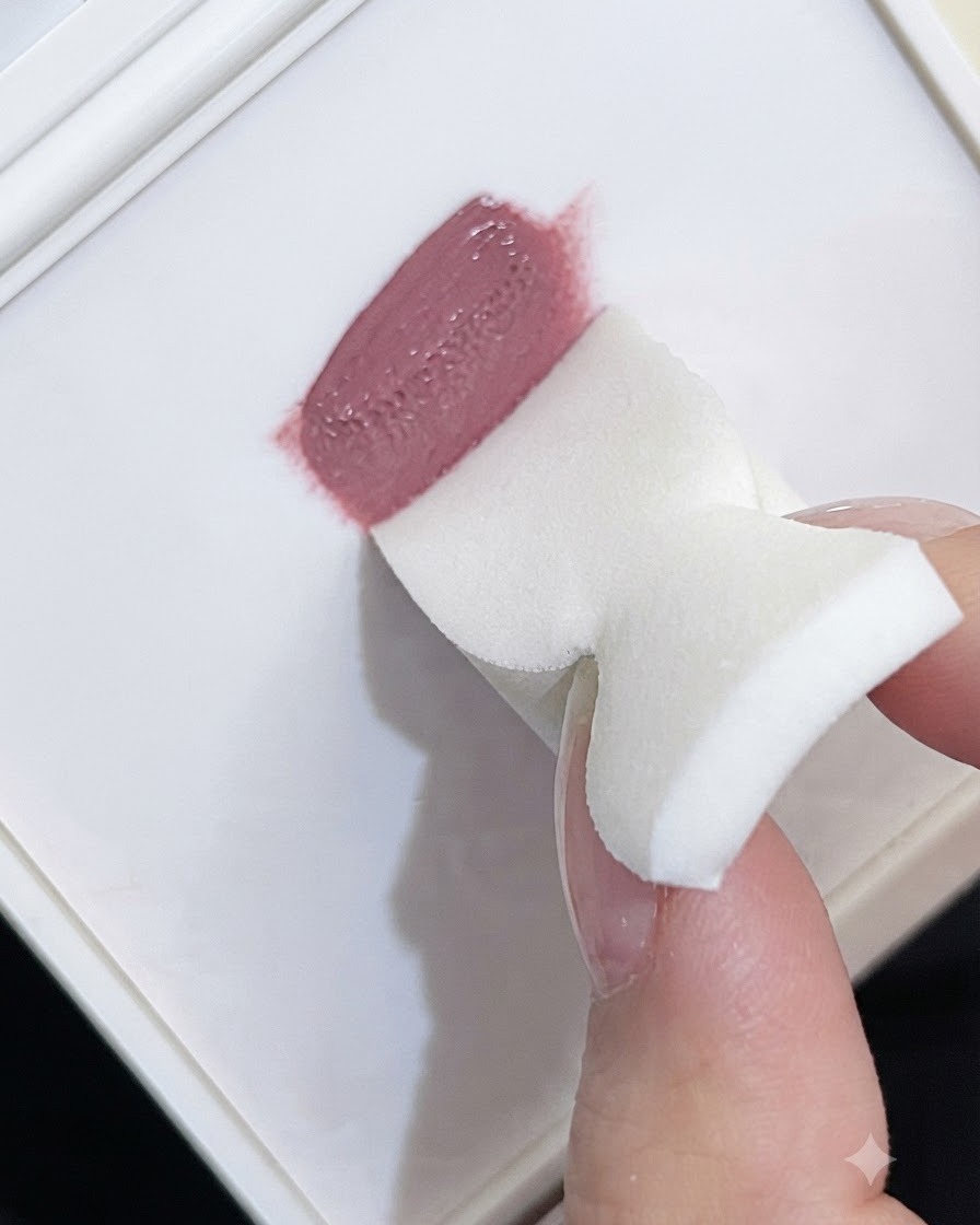 Il bordo di una spugnetta bianca che preleva gel color malva da una tavolozza.